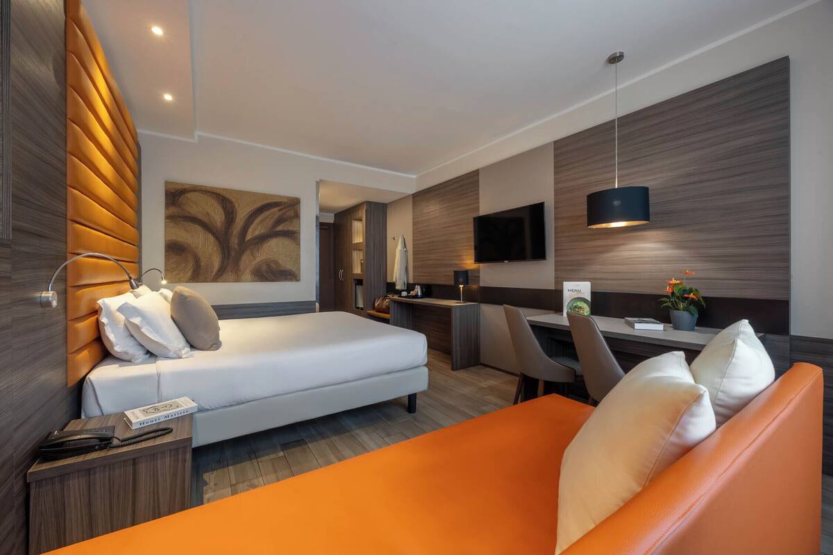A modern hotel room with a bed, a desk, a chair, a TV, and a small plant.