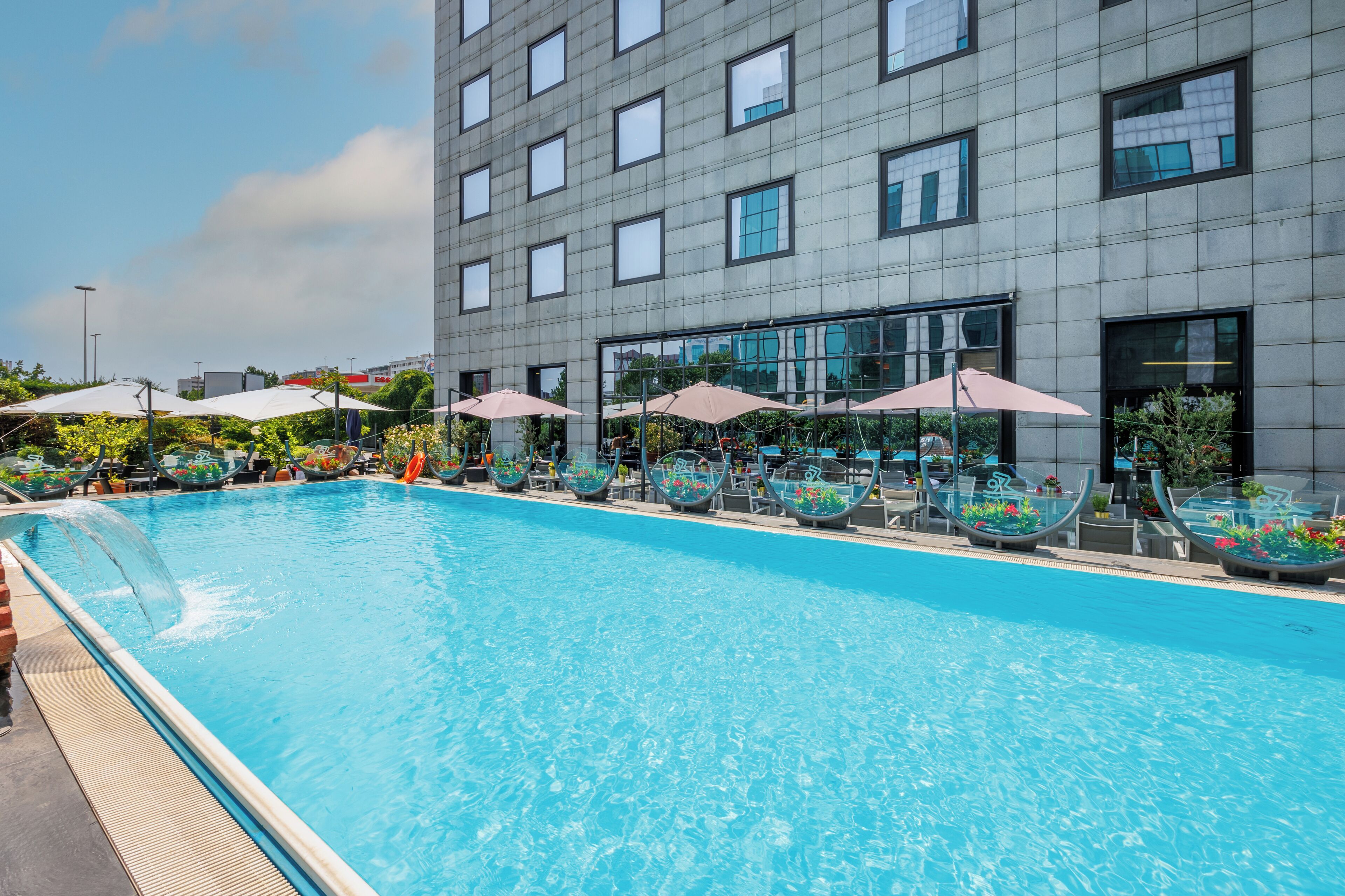 Una piscina con sillas de descanso y sombrillas, un edificio moderno con ventanas grandes, y un cielo despejado.