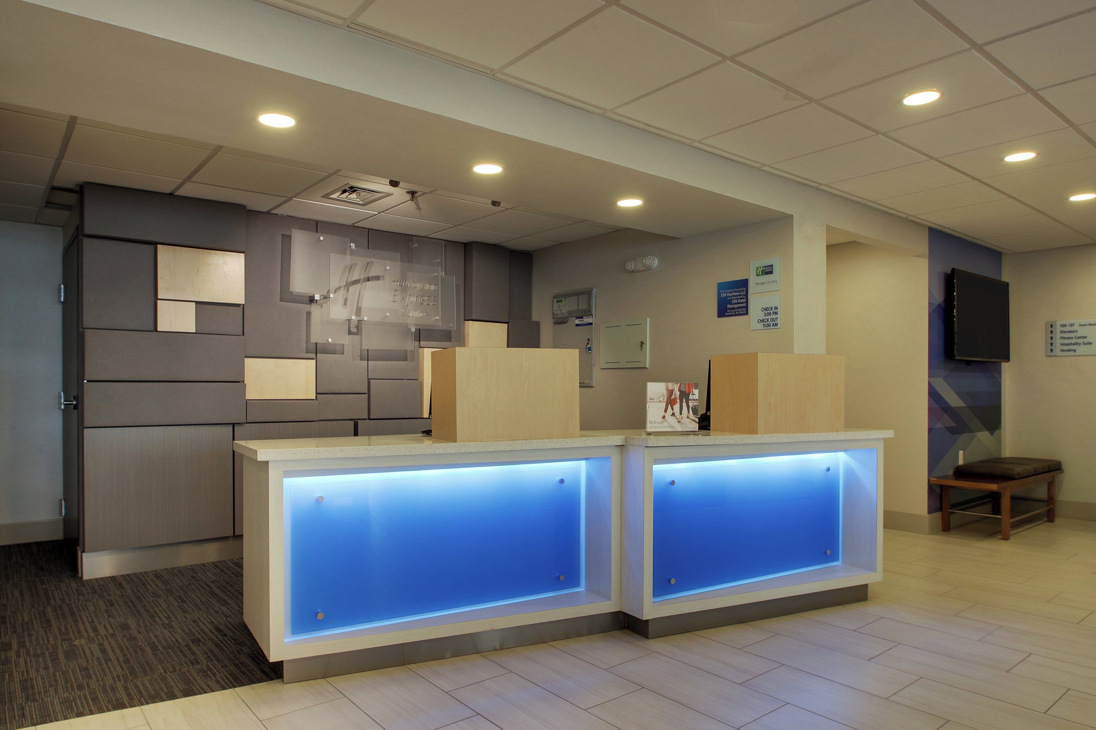 A reception area with a modern design, featuring a reception desk with illuminated blue panels, a flat-screen TV mounted on the wall, and a digital clock.