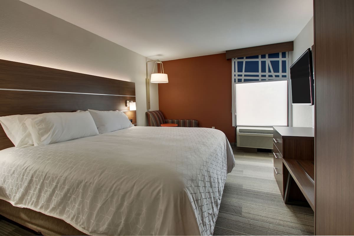 A hotel room with a large bed, a desk, a chair, a TV, and a window with blinds.