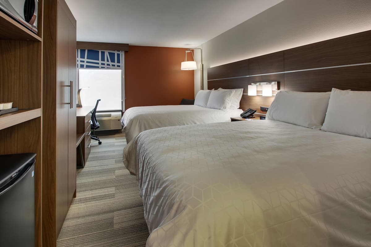 A hotel room with two beds, a desk with a chair, a lamp, and a view of the outdoors.