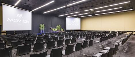 A conference room with rows of black chairs, a stage with a projector screen displaying "MOVICH," and a neutral-colored carpet.