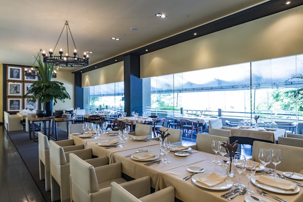 A restaurant with multiple dining tables set for guests, large windows offering a view of the outdoors, and a chandelier hanging from the ceiling.