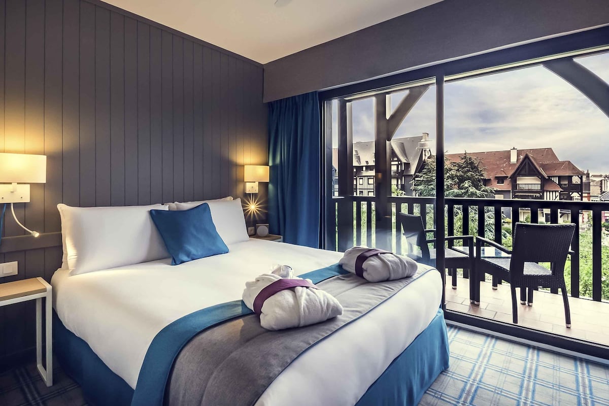 A hotel room with a large bed, two bedside lamps, a view of the balcony, and a cityscape in the background.