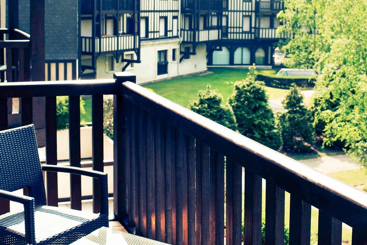 A balcony with a wicker chair, overlooking a courtyard with trees and a building with a traditional facade.