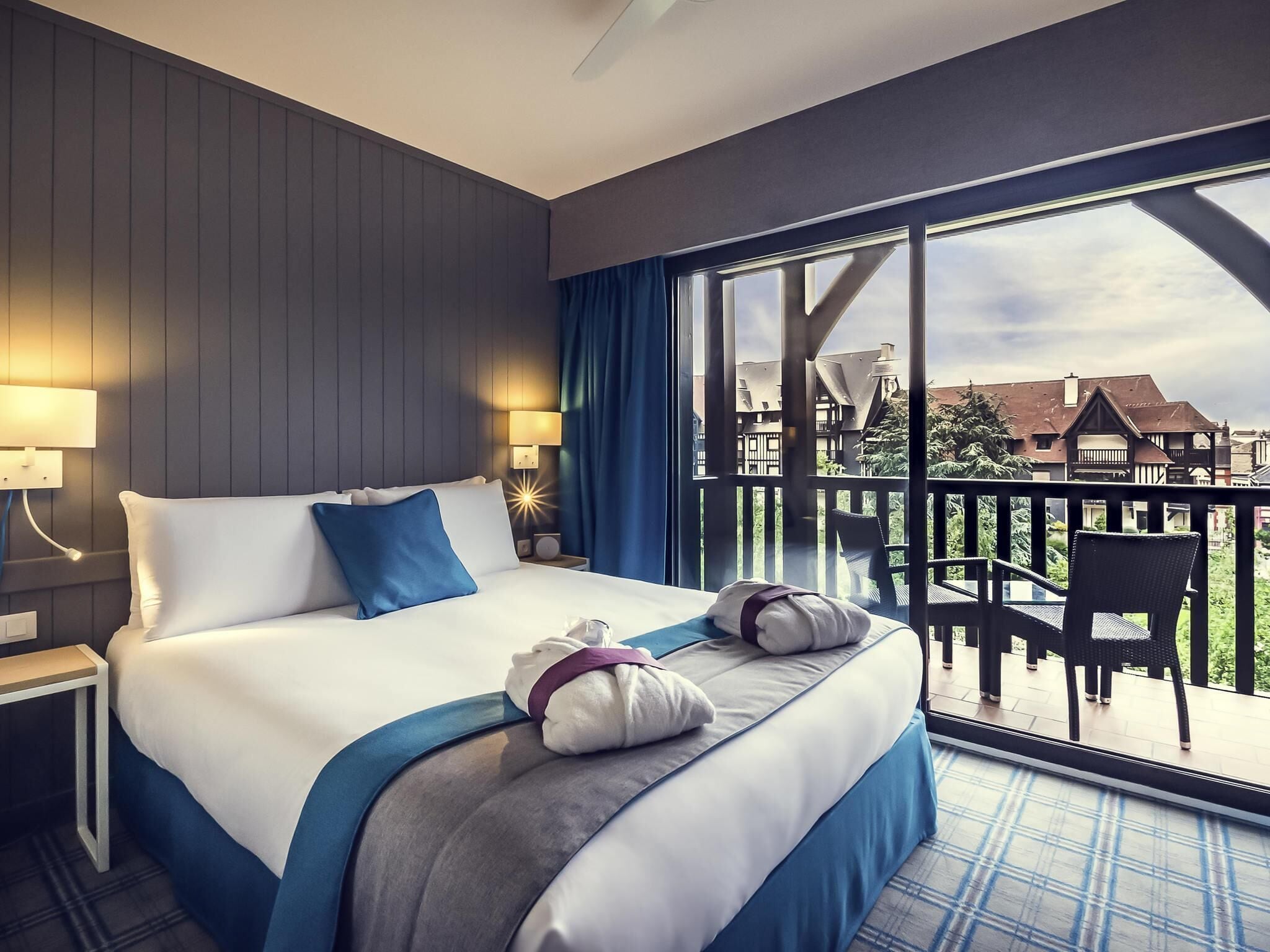 A hotel room with a large bed, two bedside lamps, a view of buildings through a balcony.