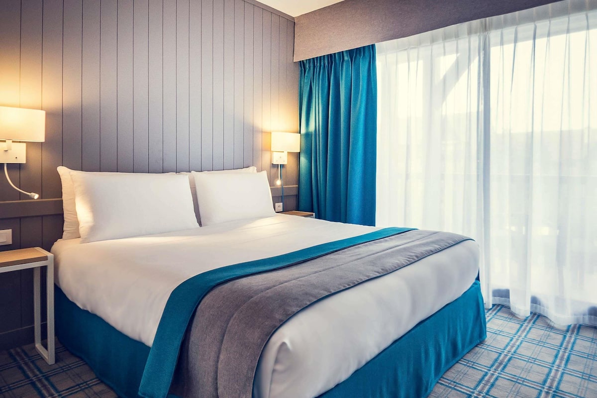 A hotel room with a large bed, two bedside tables, a nightstand, a window with curtains, and a wall-mounted lamp.