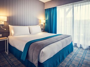A hotel room with a bed, nightstands, lamps, a window with curtains, and a plaid-patterned carpet. - Mercure Deauville Centre (Deauville)