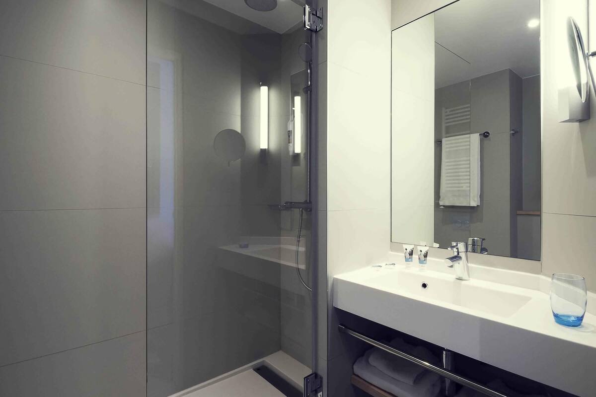 Une salle de bain moderne avec une douche à parois de verre, un grand miroir et un lavabo blanc avec un verre d’eau.