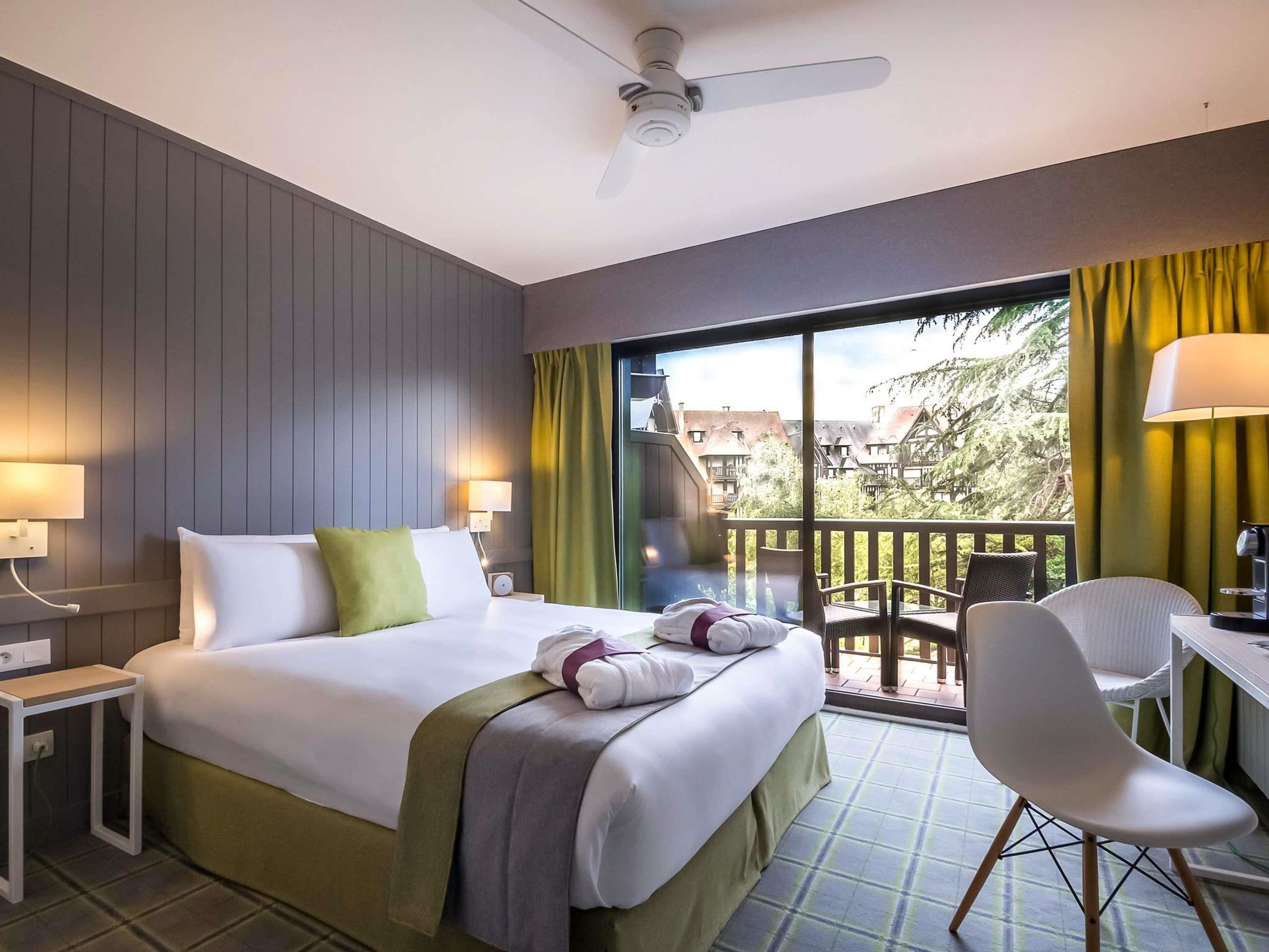 A hotel room with a bed, a desk, a chair, a balcony with a view, and a ceiling fan.