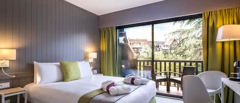 A hotel room with a bed, a desk, a chair, a balcony with a view, and a ceiling fan.