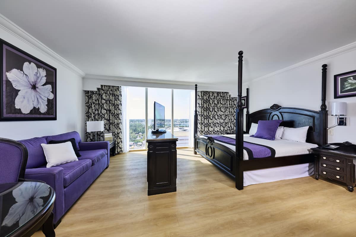 A modern hotel room with a large bed, a sofa, a small table, and a view of the city through large windows.