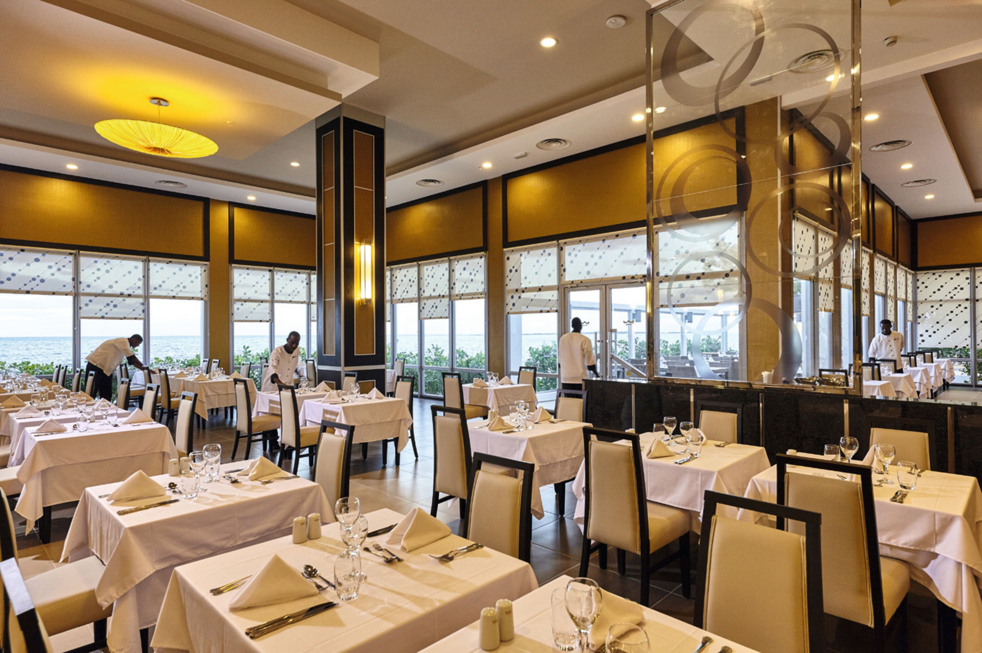 A restaurant with tables set for dining, large windows, and a view of the sea.