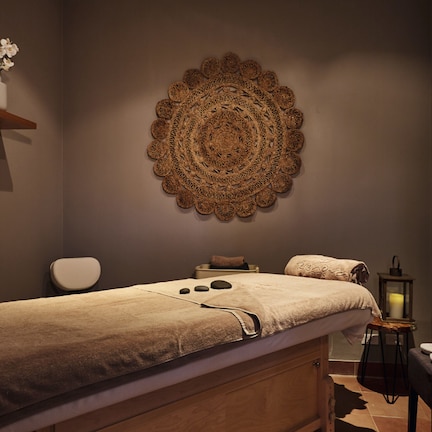 A spa room with a massage table, a wall-mounted decorative piece, a shelf with candles, and a small table with a candle.