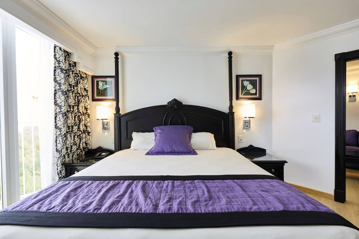 A four-poster bed with a purple accent pillow, white bedding, and two bedside lamps.