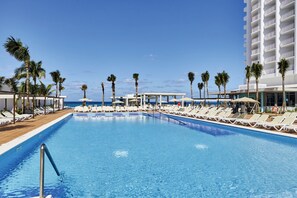 Área de piscina com espreguiçadeiras e guarda-sóis, palmeiras e um edifício com varandas.
