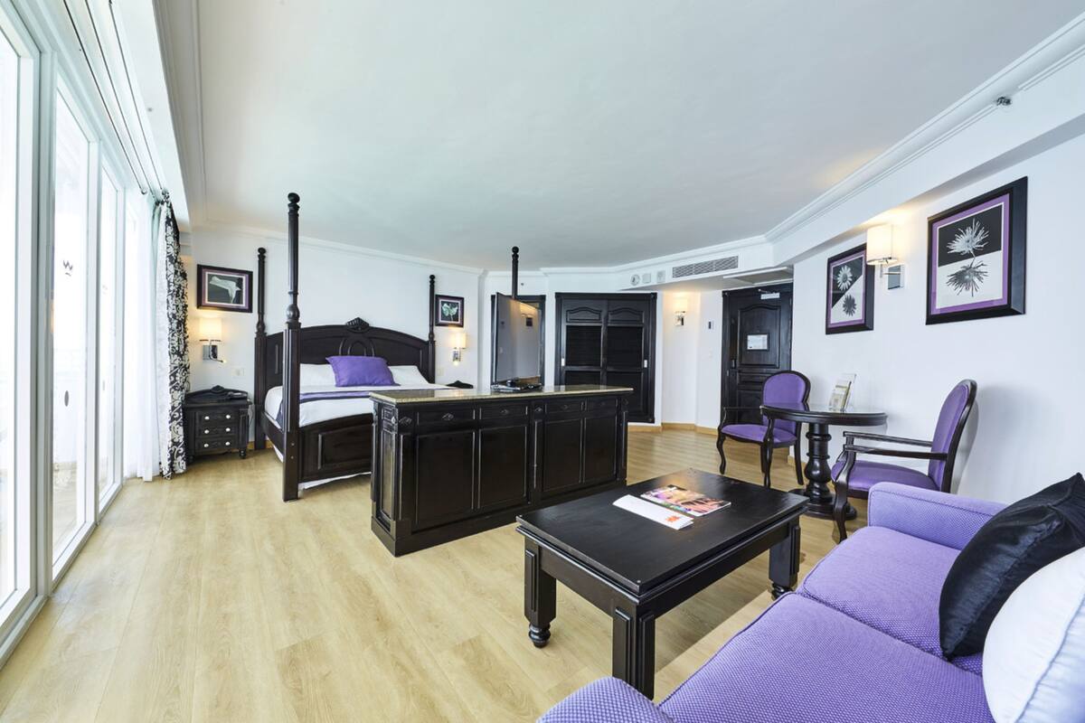 A modern hotel room with a large bed, a sofa, a coffee table, and a dining area.