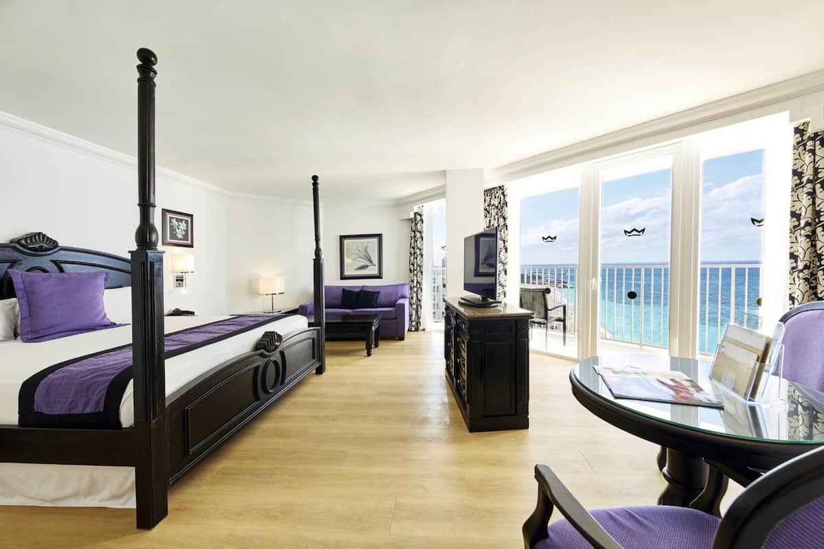 A bedroom with a four-poster bed, a sofa, a small table, and a balcony with a view of the sea.
