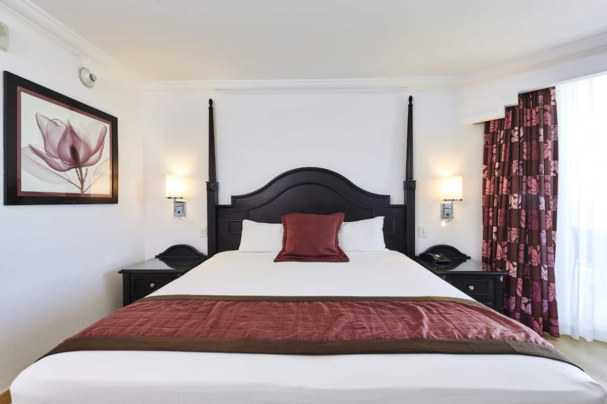 A neatly made bed with a dark headboard, two bedside tables with lamps, a framed floral artwork on the wall, and a window with curtains.