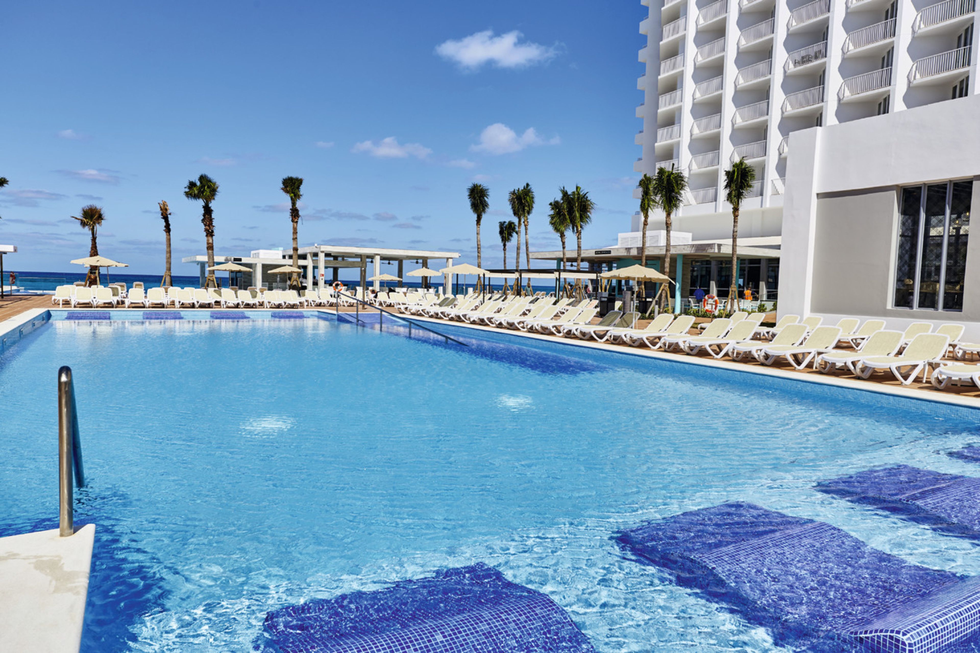 A swimming pool with lounge chairs and umbrellas, a building with balconies, and palm trees.