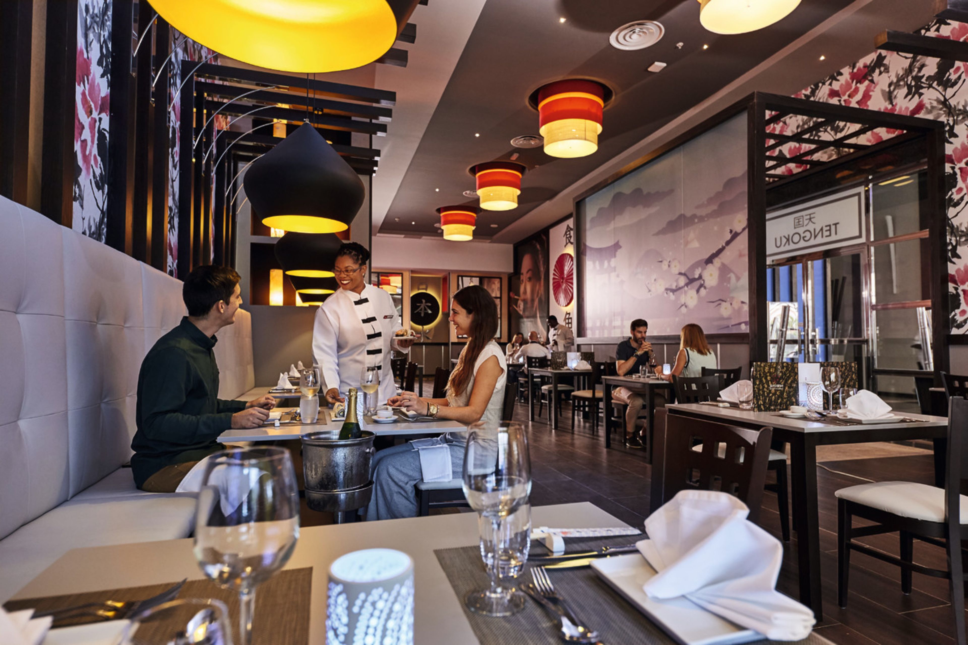 A restaurant interior with multiple dining tables, hanging pendant lights, and a large mural on the wall.
