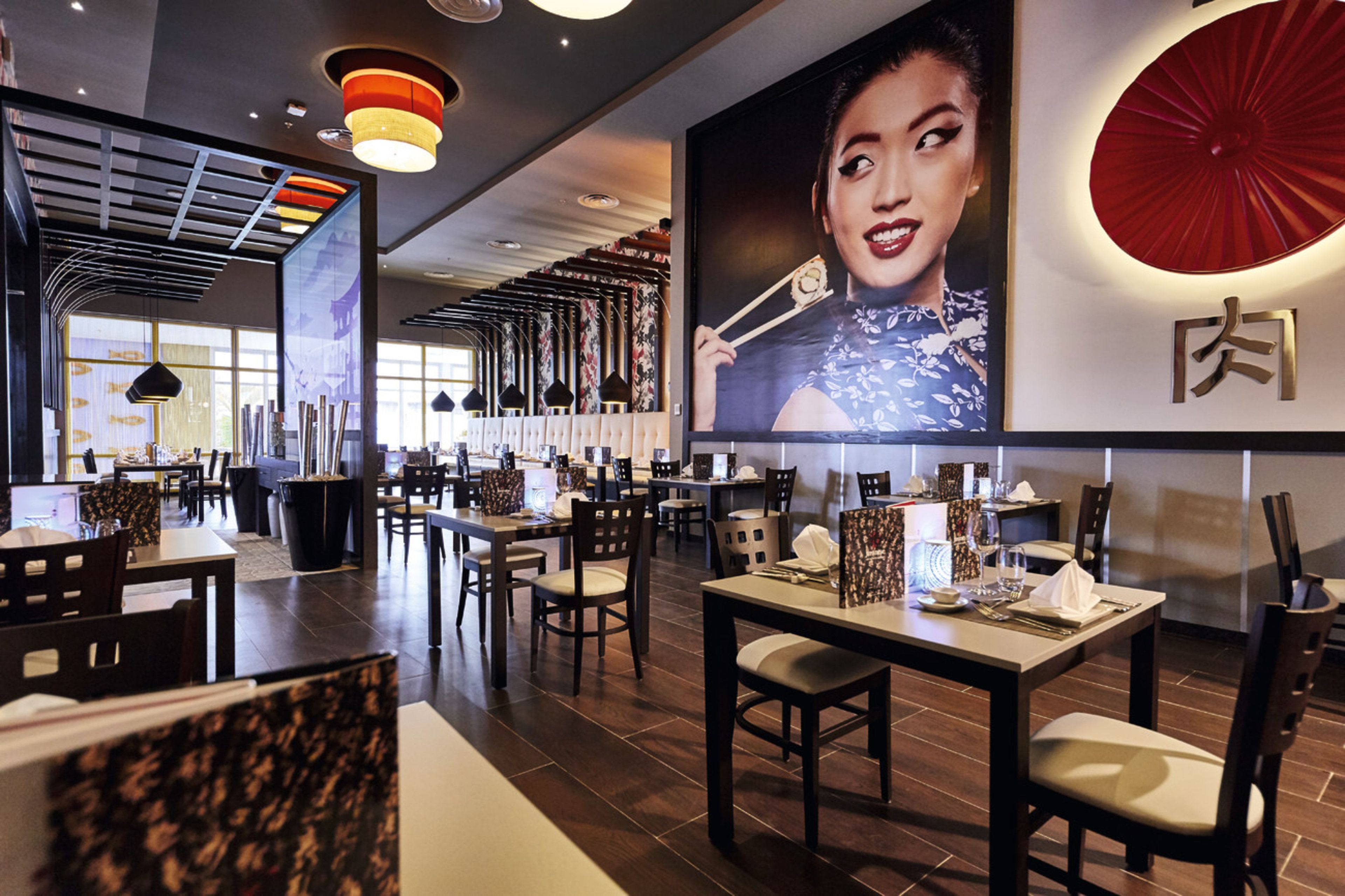 A restaurant interior with multiple dining tables and chairs, a large wall-mounted artwork, and a ceiling with exposed beams and lighting fixtures.