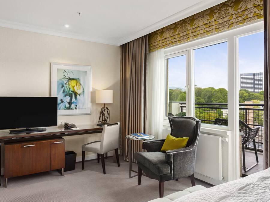 A hotel room with a large window, a desk with a flat-screen TV, a comfortable armchair, and a bed.