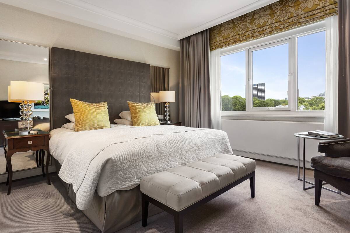 A hotel room with a large bed, two bedside lamps, a bench, a small table, and a view of the city through large windows.