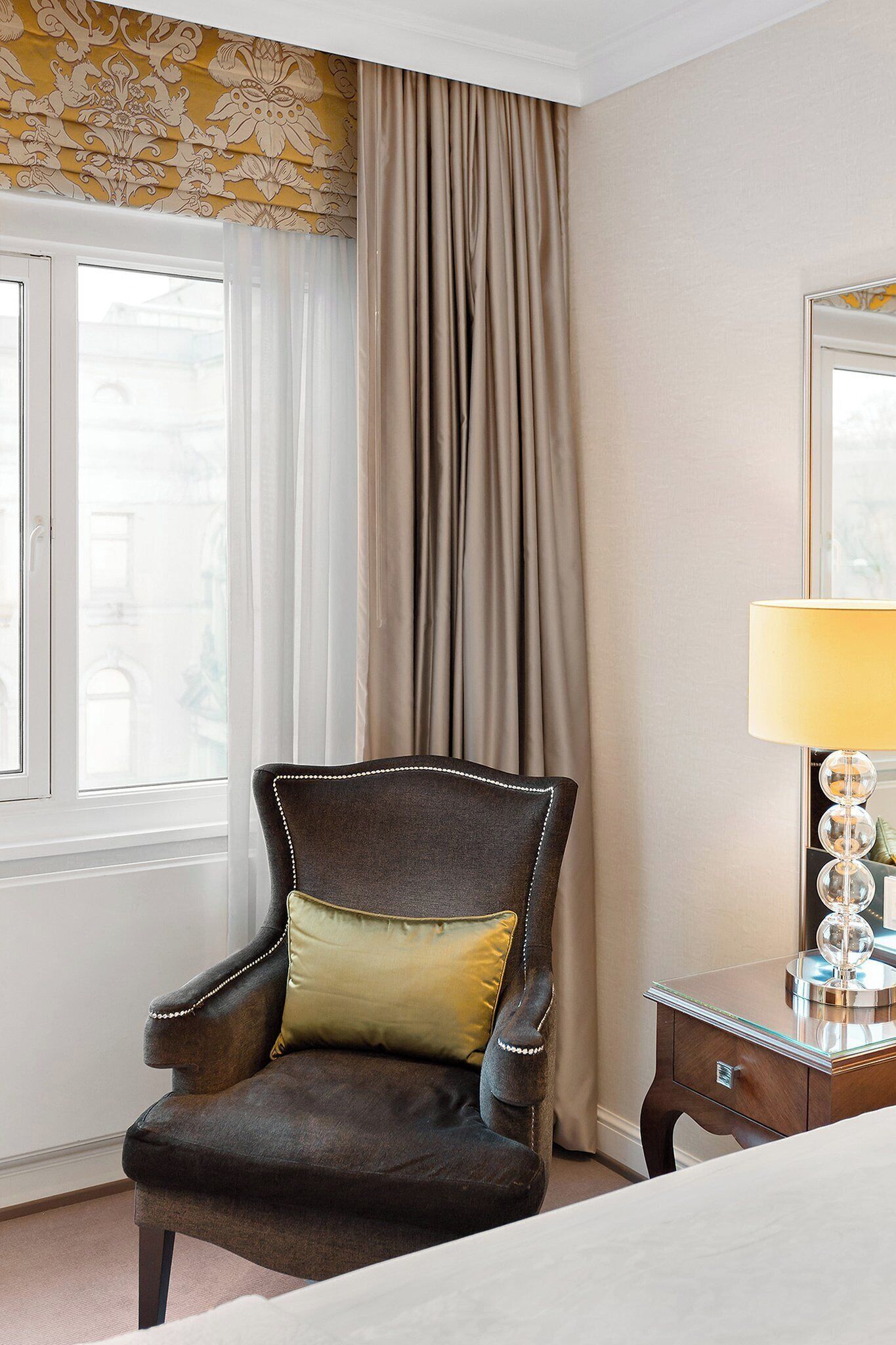 A hotel room with a dark leather armchair, a beige curtain, a glass side table with a lamp, and a window with a view of buildings.