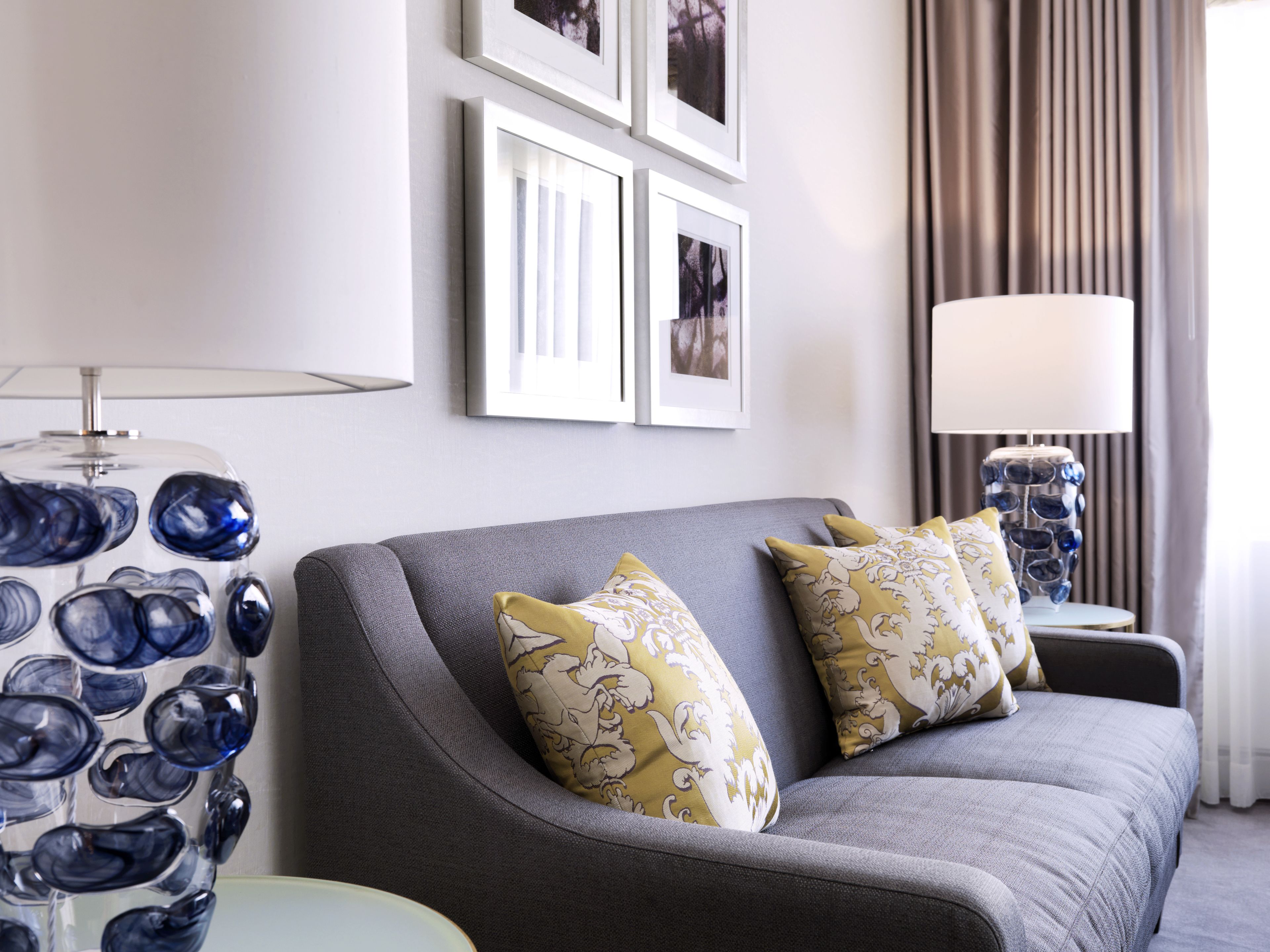 A modern living room with a grey sofa, two patterned cushions, a glass lamp, and framed pictures on the wall.