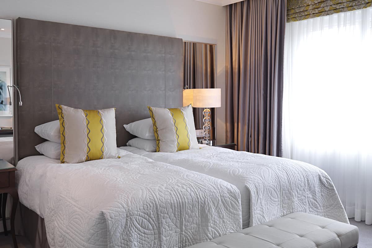 A hotel room with two beds, a large headboard, a bedside table with a lamp, and a window with curtains.