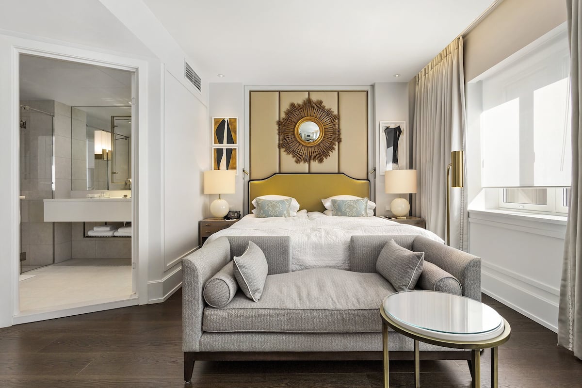 A modern hotel room with a large bed, a sofa, a small table, and a bathroom visible through an open door.