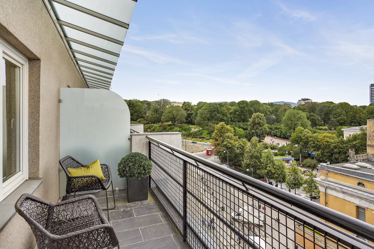 A balcony with wicker chairs and a potted plant, overlooking a cityscape with trees and buildings.