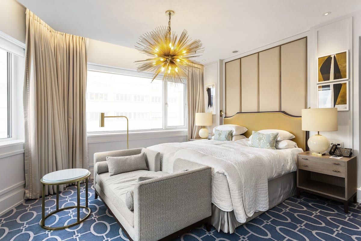 A hotel room with a large bed, a bedside table with a lamp, a small round table, and a modern chandelier.