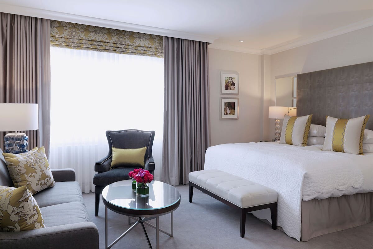 A hotel room with a bed, a sofa, a chair, a small table with a vase of flowers, and a window with curtains.