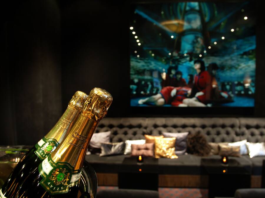 En flaska champagne i ett svagt upplyst rum, där en stor skärm visar en scen med människor.