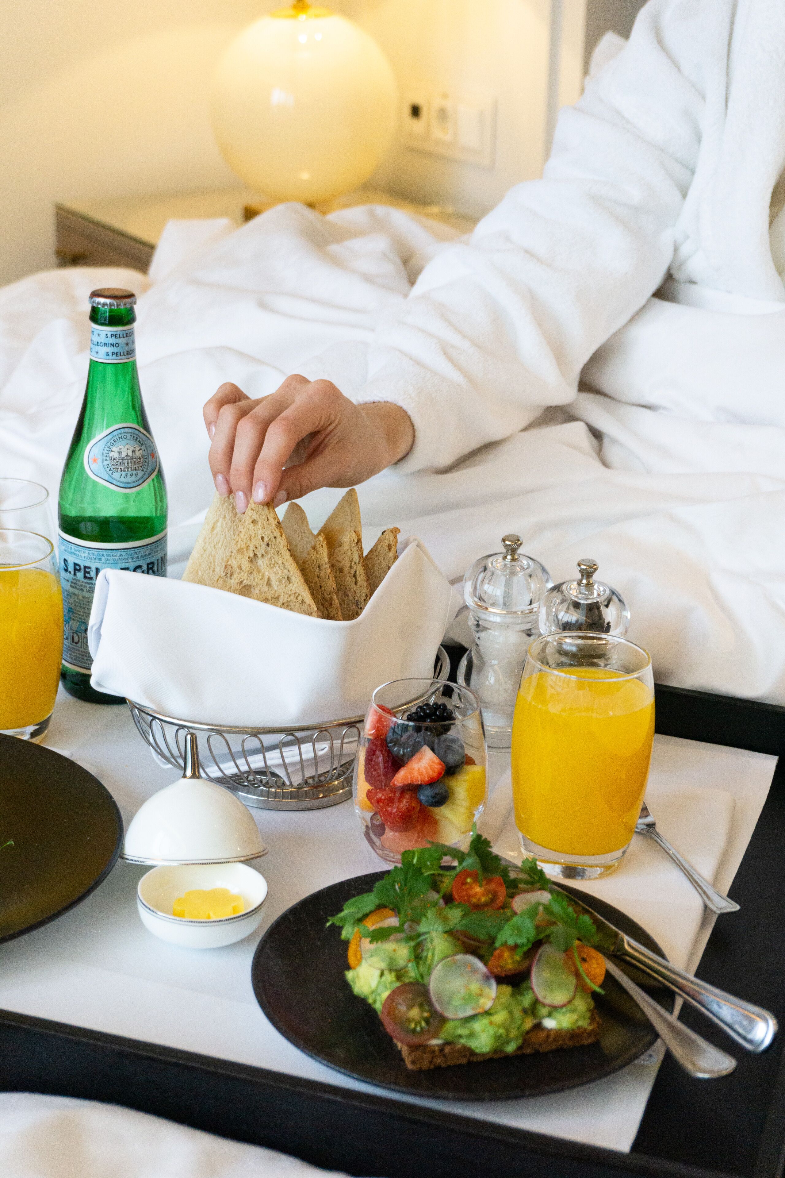 A breakfast tray with toast, avocado, berries, and orange juice on a bed.
