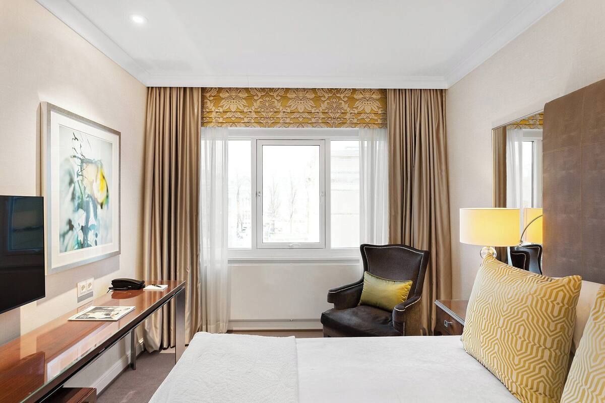 A hotel room with a bed, a desk with a chair, a television, and a window with curtains.