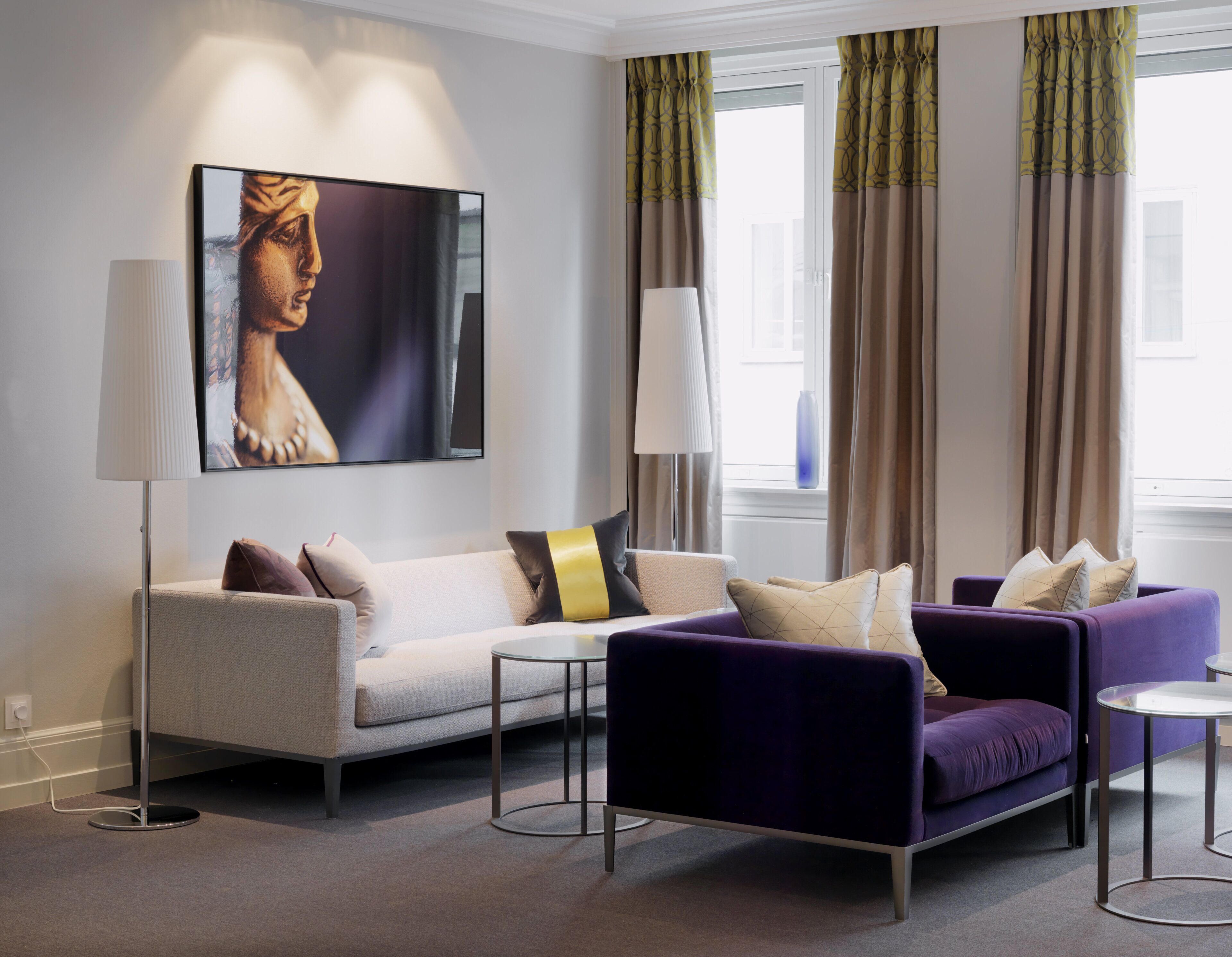A modern living room with a sofa, armchairs, and a coffee table. There is a painting on the wall and a lamp beside the sofa.