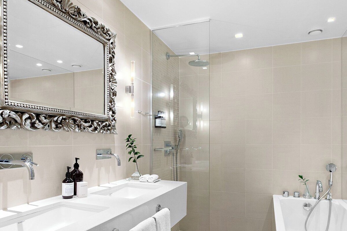 A modern bathroom with a large mirror, two sinks, a shower, and a toilet.