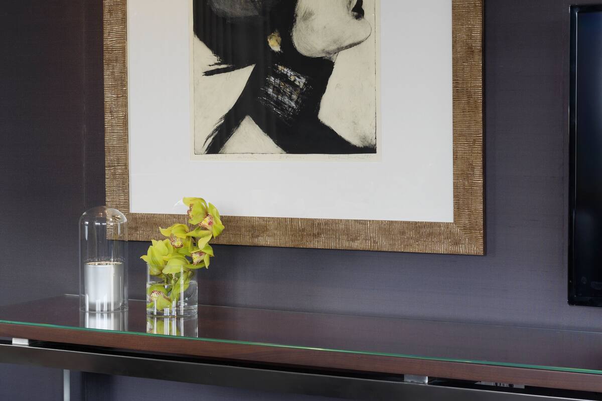A framed artwork on a wall, a glass vase with flowers, and two stools.