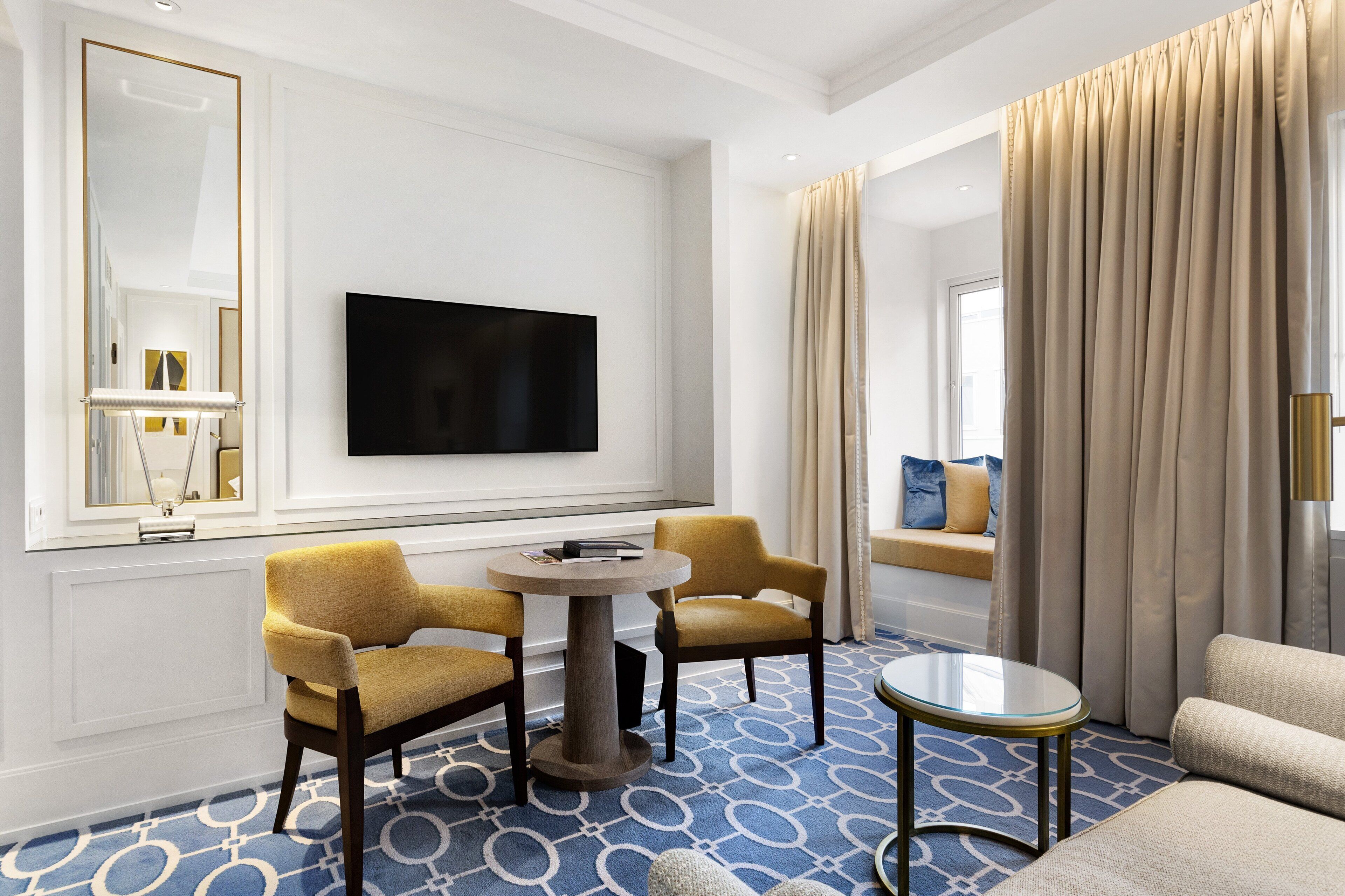 A hotel room with a flat-screen TV mounted on the wall, two yellow armchairs around a round table, a sofa, and a large window with beige curtains.