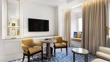 A hotel room with a flat-screen TV mounted on the wall, two yellow armchairs around a round table, a sofa, and a large window with beige curtains.