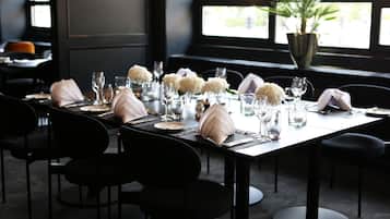 A dining table set with white napkins, plates, and glasses, surrounded by black chairs, in a room with large windows and a potted plant.