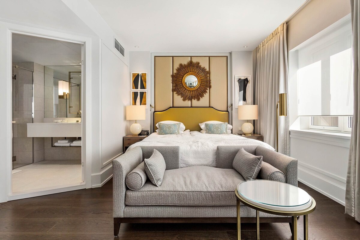 Habitación de hotel con cama, sofá, mesa redonda, espejo y vistas al exterior.