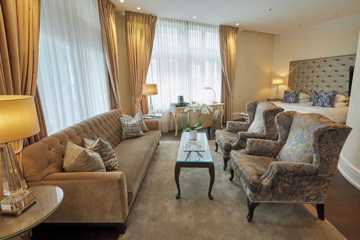 A well-appointed hotel room with a sofa, armchairs, a coffee table, and a bed with a tufted headboard.