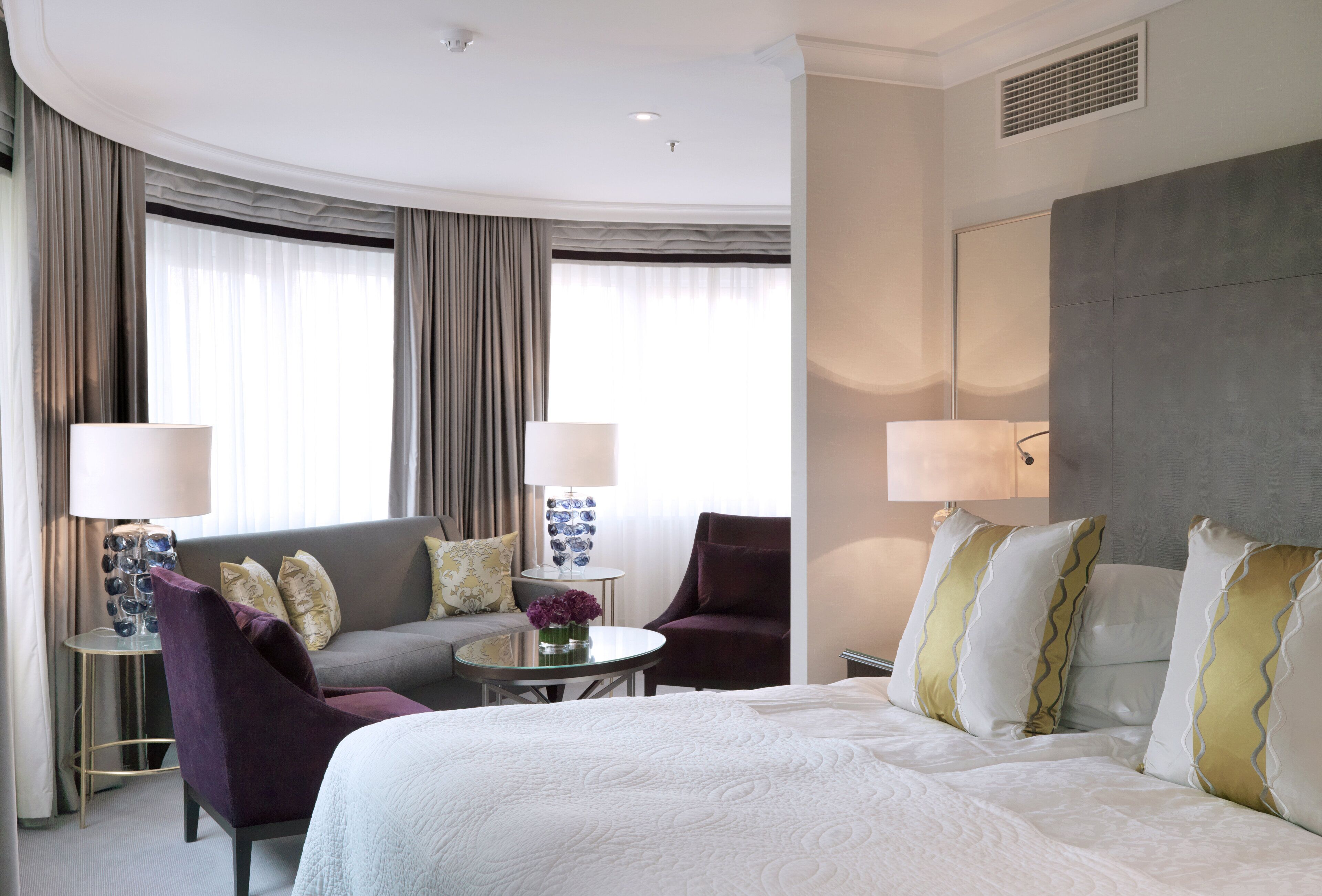 A hotel room with a large bed, a sofa, a chair, a small table with a lamp, and a vase with flowers.