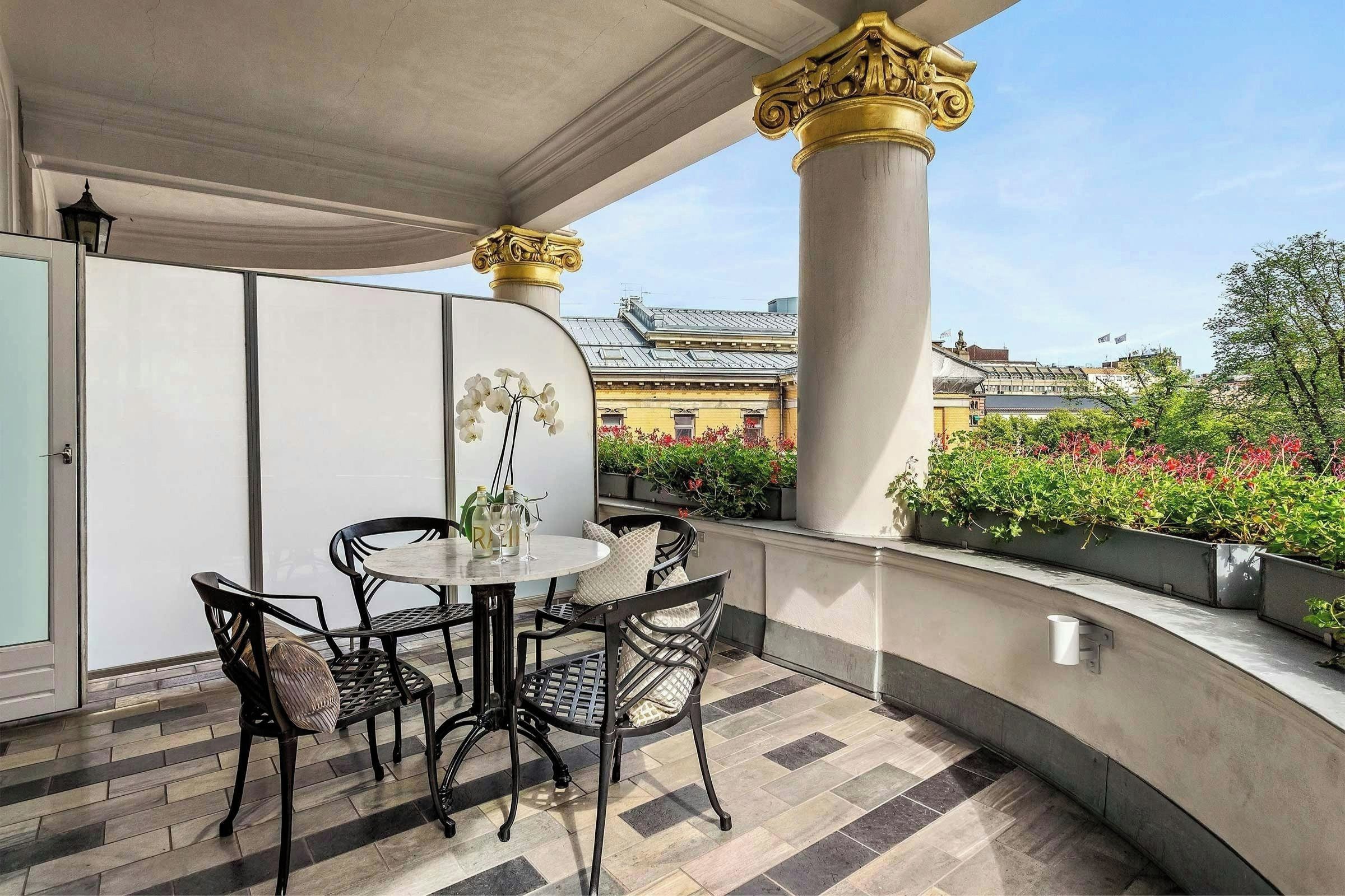 Un balcon avec une table ronde et des chaises, un pot d’orchidée et une vue sur des bâtiments et de la verdure.