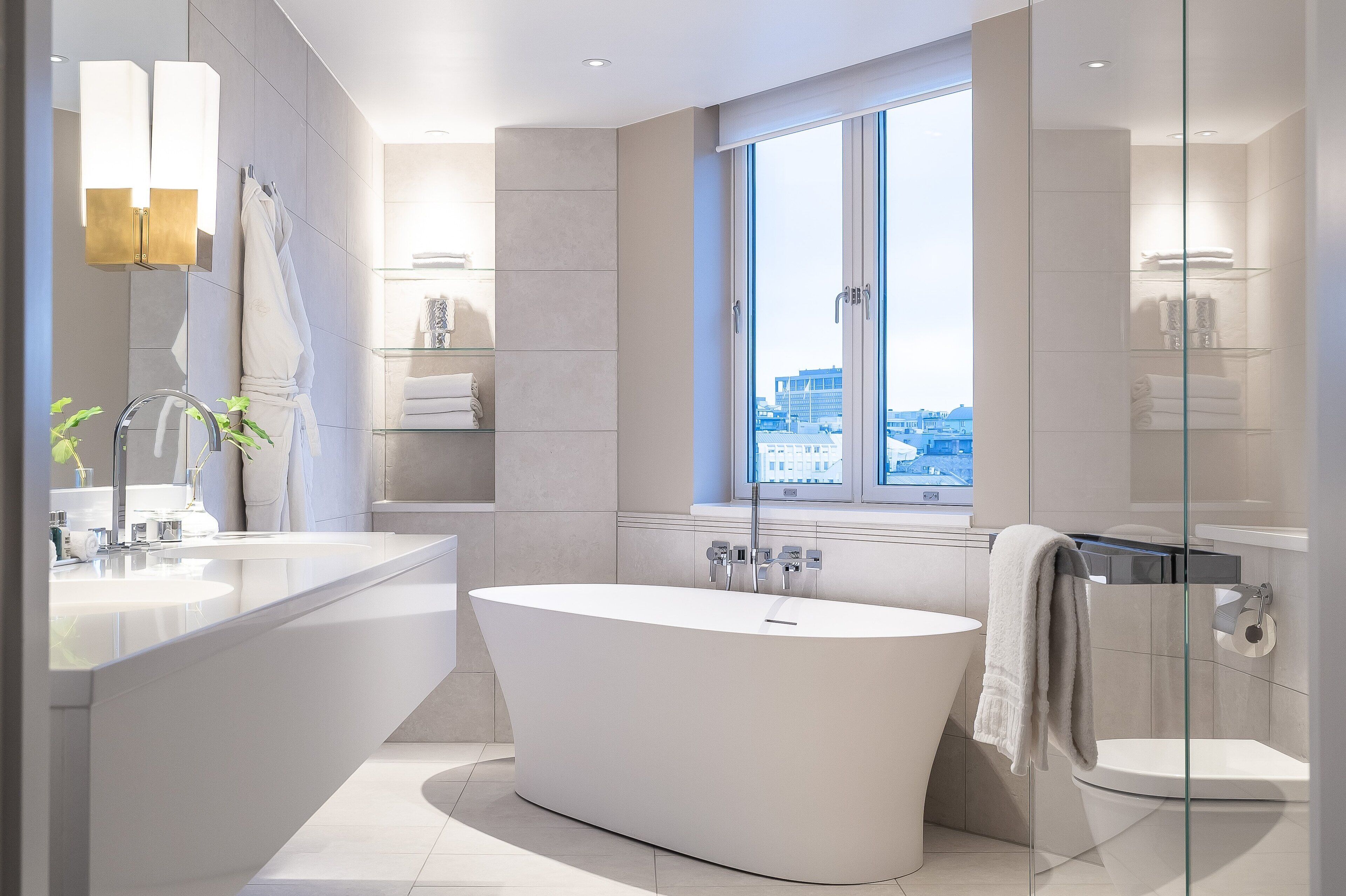 A modern bathroom with a freestanding bathtub, a large mirror, and a window offering a city view.