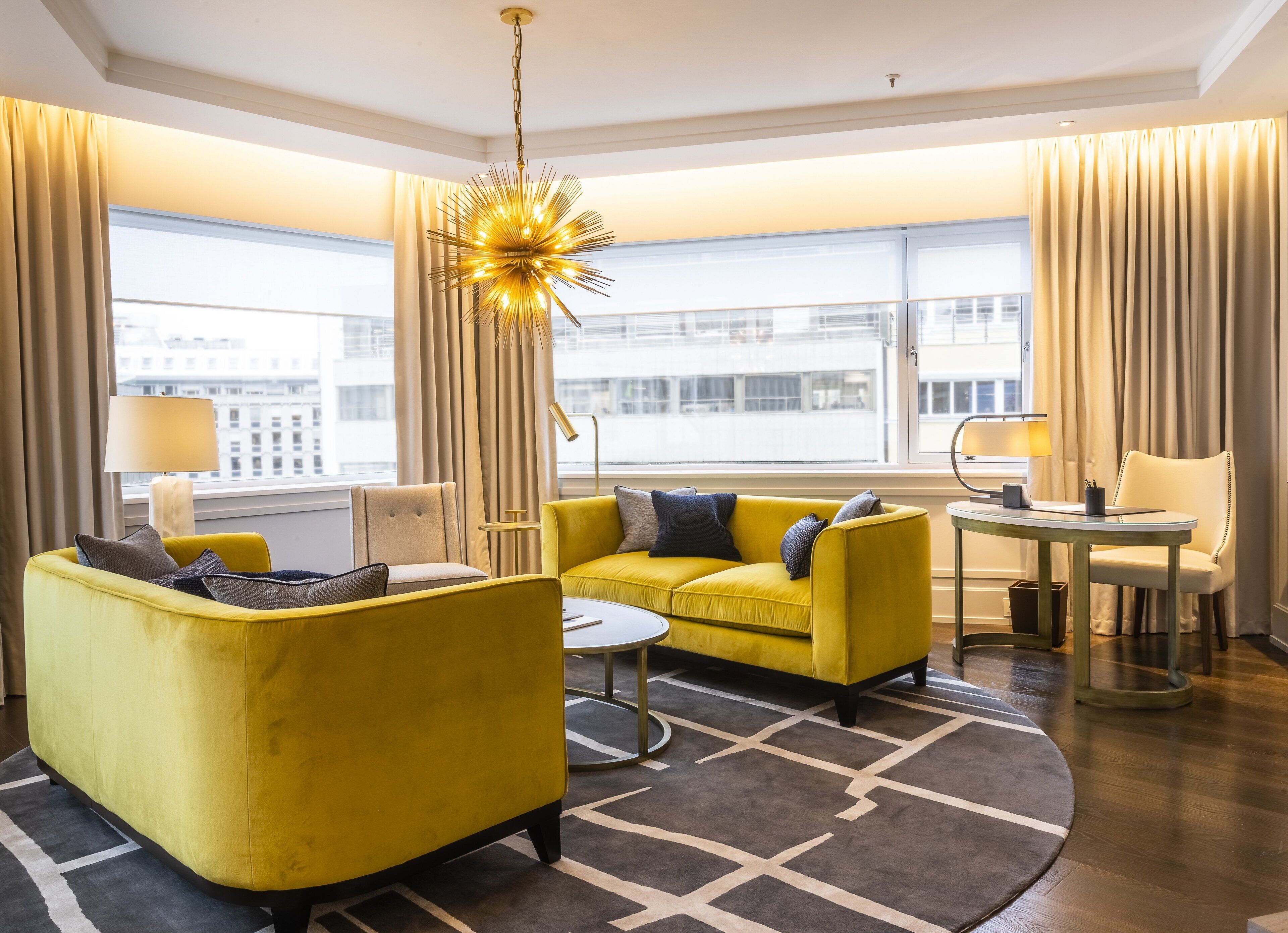 Una camera d'albergo moderna con un divano giallo, un tavolino rotondo e un lampadario. Grandi finestre con tende, una lampada e vista sugli edifici.